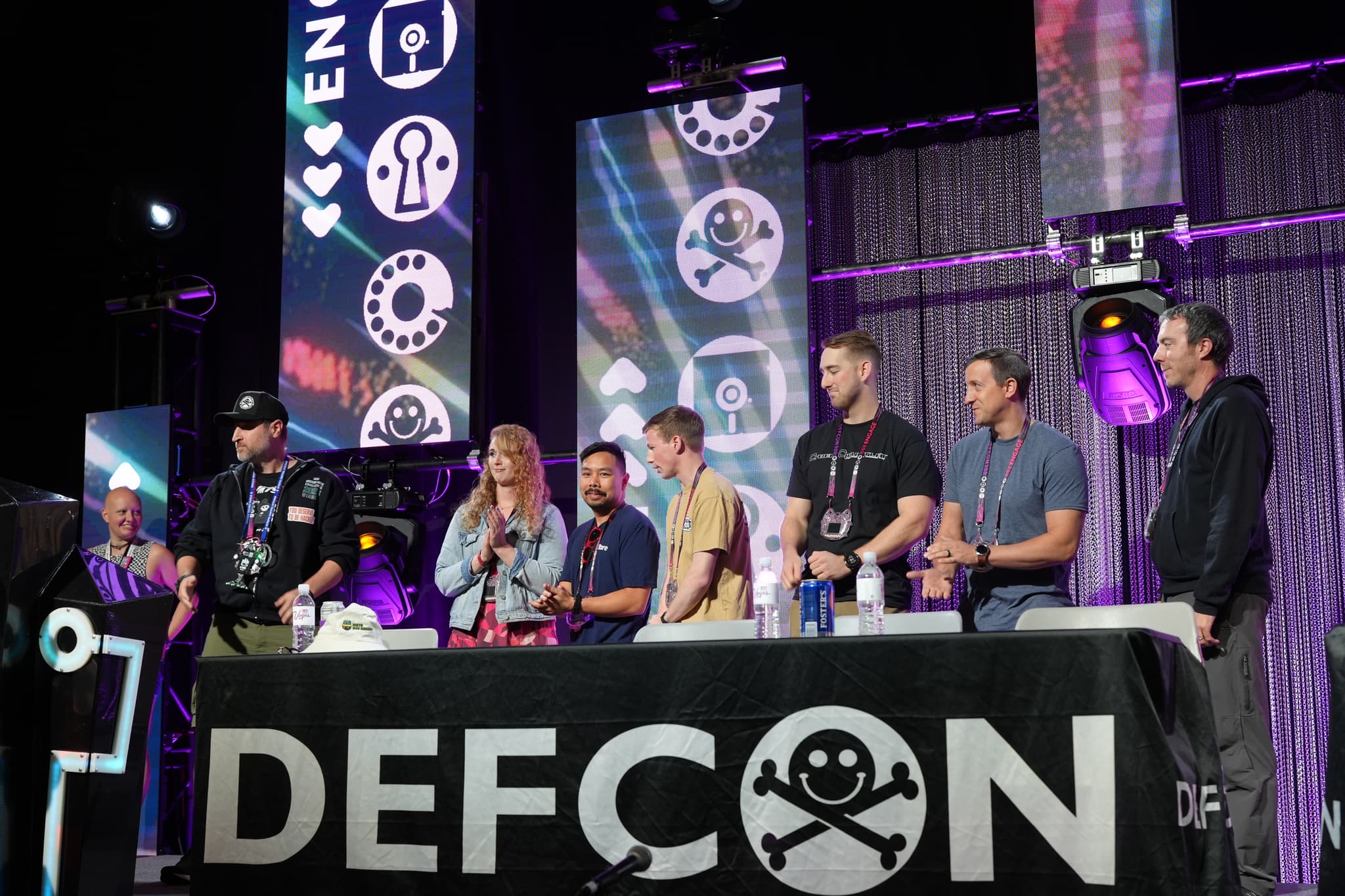 A group of people standing behind a table with a DEFCON banner, with various tech-themed banners and lighting in the background