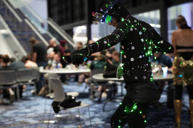 A person wearing a motion capture suit with reflective markers, surrounded by people sitting at tables in a conference or event setting