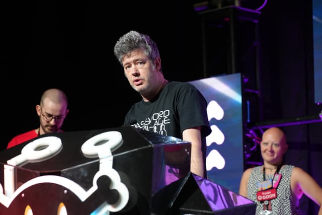 Three people on a stage, with one person speaking at a podium decorated with a stylized design. The background features colorful lighting and geometric shapes