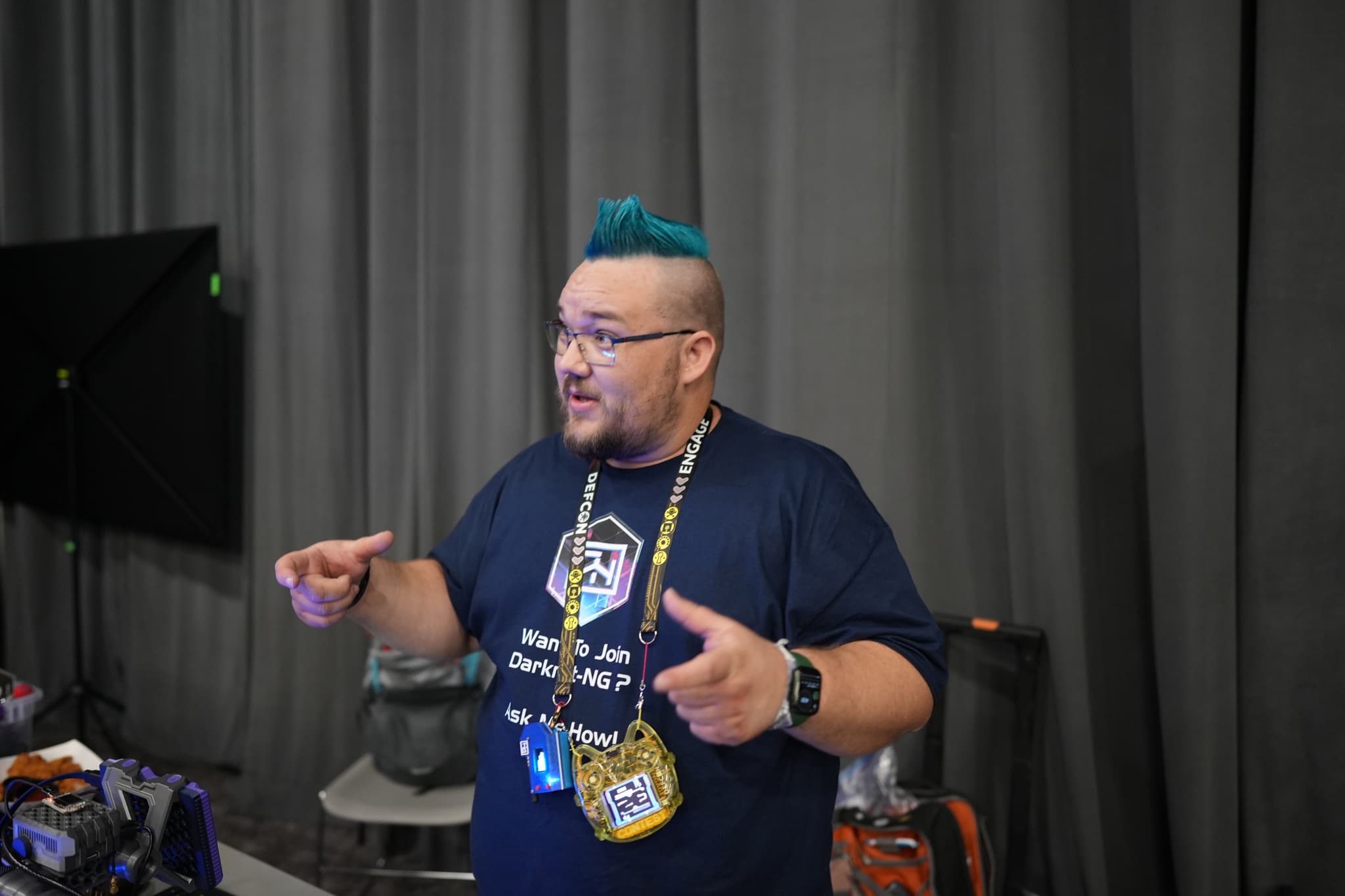 A person with a blue mohawk and glasses is gesturing with their hands while wearing a dark t-shirt and a lanyard with badges. They are standing in front of a gray curtain with some equipment and chairs in the background