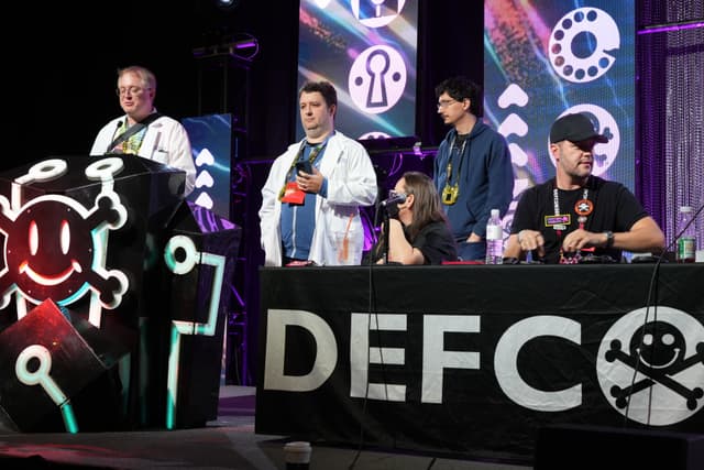 A group of people on a stage at DEFCON, with one person standing at a podium decorated with a skull and crossbones symbol, and others seated or standing behind a table with the DEFCON logo. The background features various tech-themed graphics