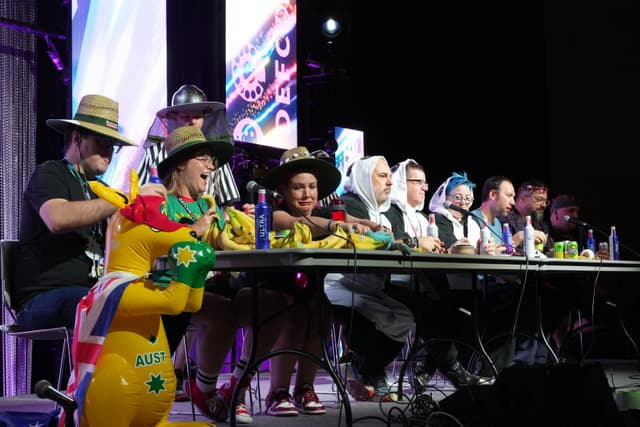 A group of people sitting at a long table, wearing various costumes and hats, with colorful decorations and props in front of them