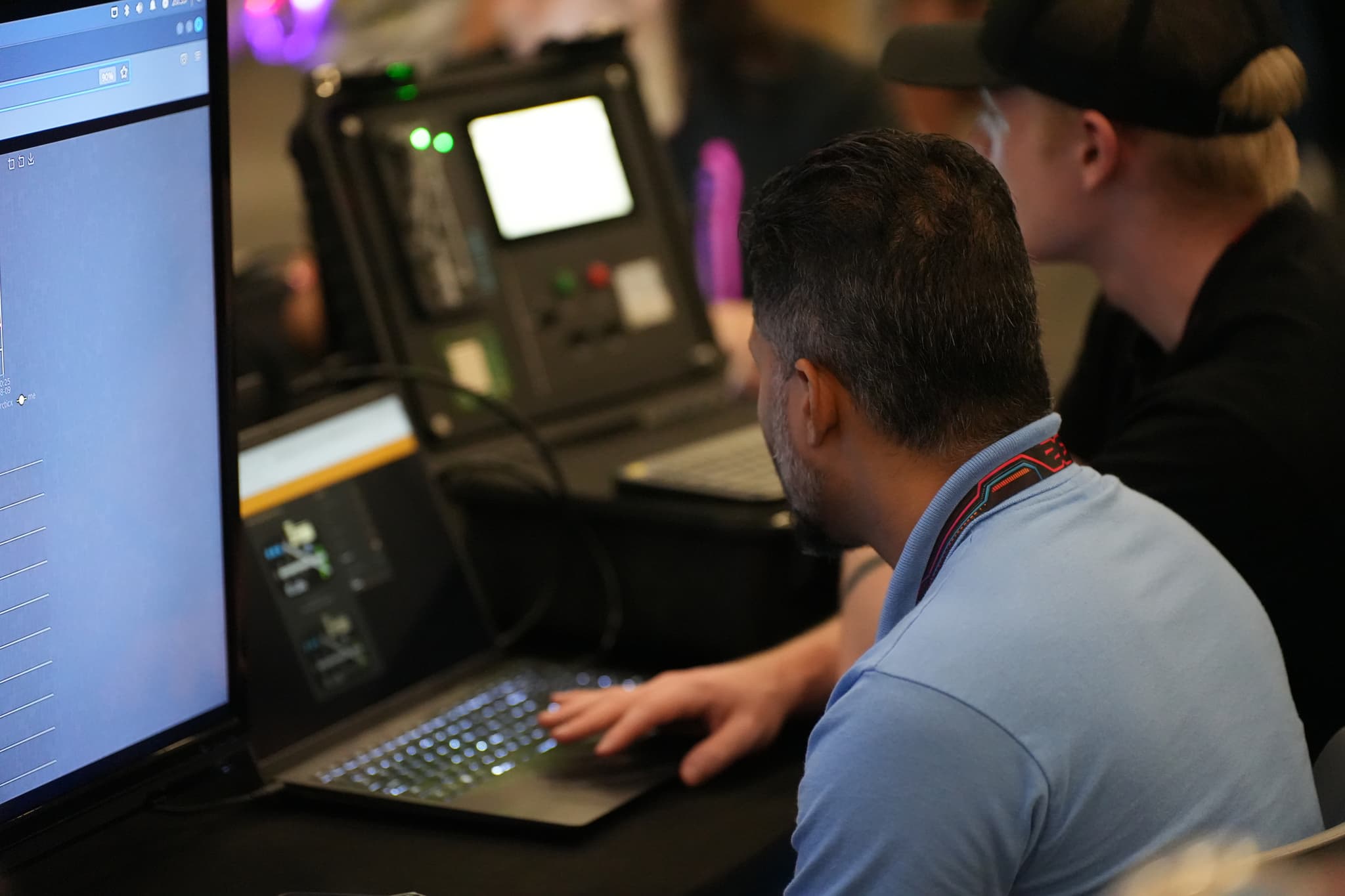 Two individuals working on laptops, with one person wearing a light blue shirt and the other in a black shirt and cap, in a dimly lit environment with electronic equipment in the background