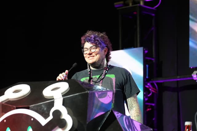 A person with glasses and curly hair stands behind a podium, speaking into a microphone. The podium has a design featuring a stylized character with large eyes. The background includes stage lighting and screens