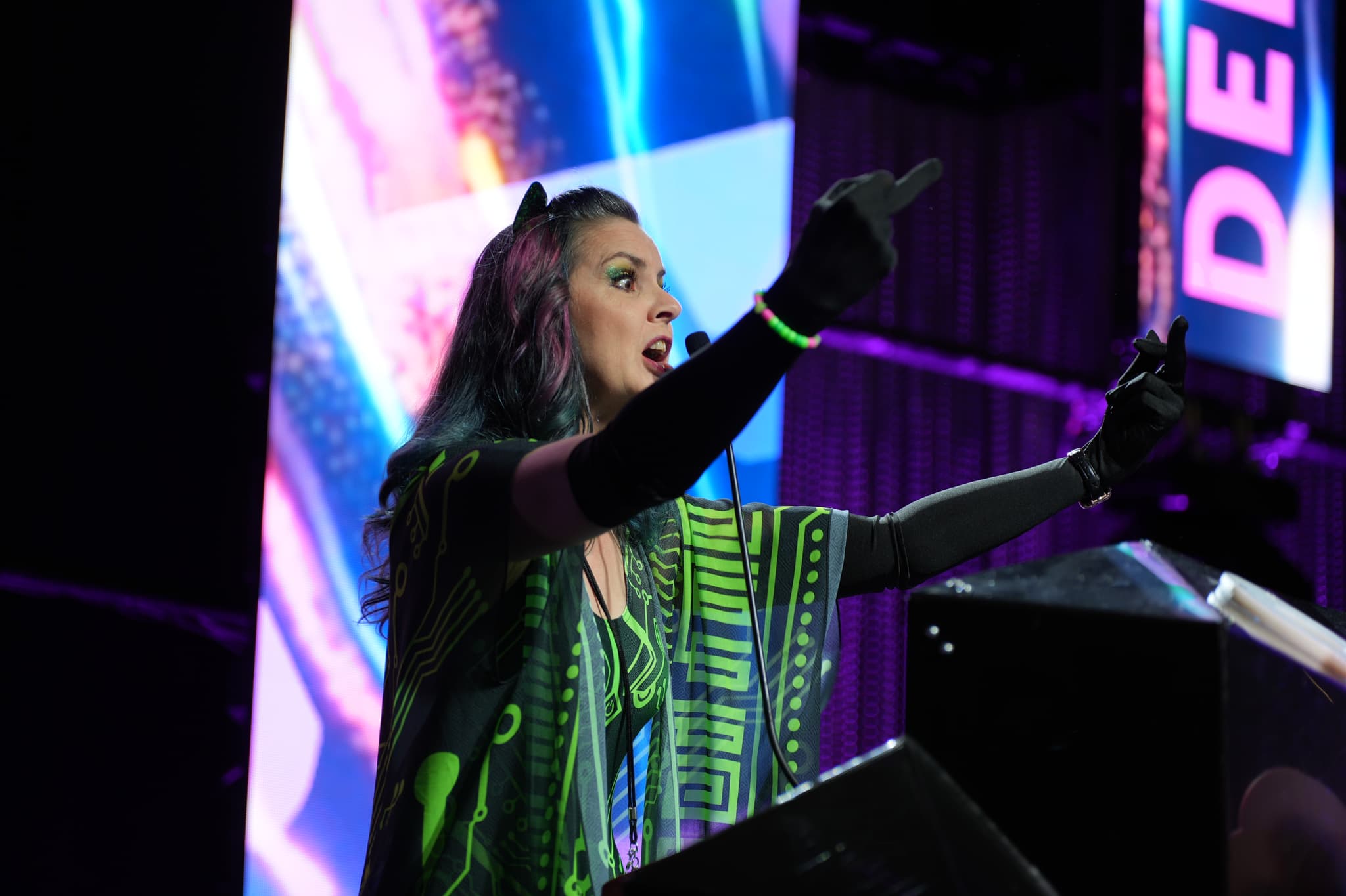 A person with long hair and wearing a colorful outfit is speaking passionately at a podium, with vibrant lights and screens in the background