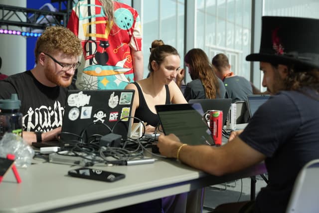 A group of people working on laptops at a table, with various stickers on the laptops and a can of soda nearby