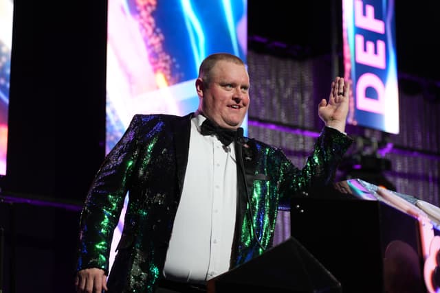 A man in a shiny, sequined jacket and bow tie stands on a stage, raising his right hand