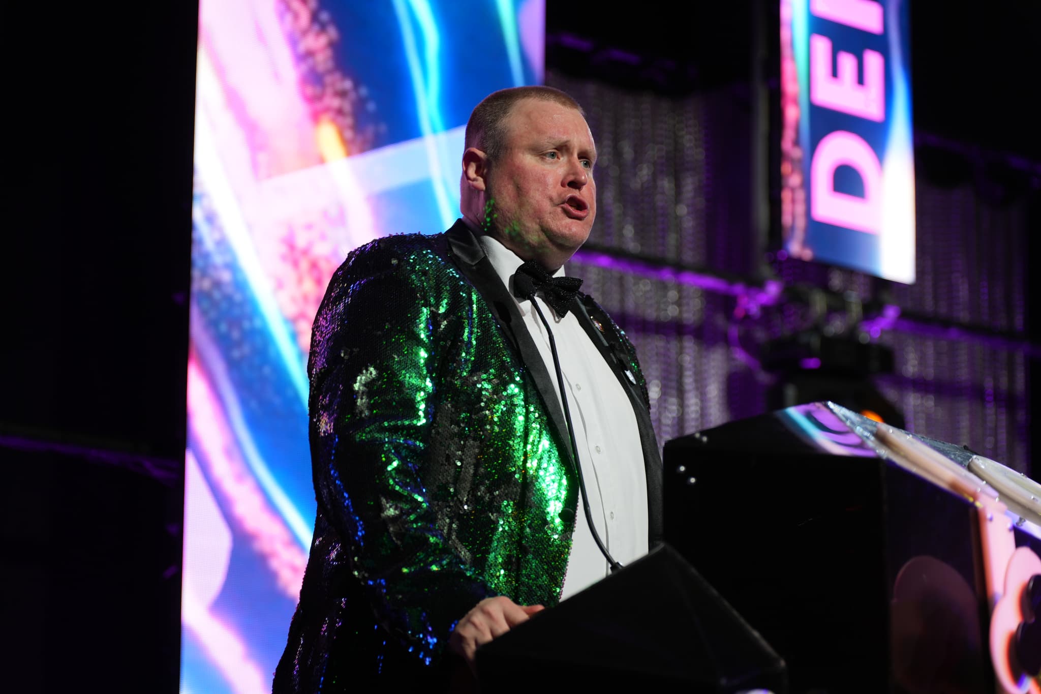 A man in a shiny green jacket and bow tie is speaking at a podium, with colorful lights and a DE sign in the background