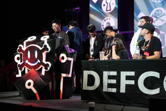 A group of people gathered around a table with a DEFCON sign, featuring a large emblem with a smiling skull and crossbones