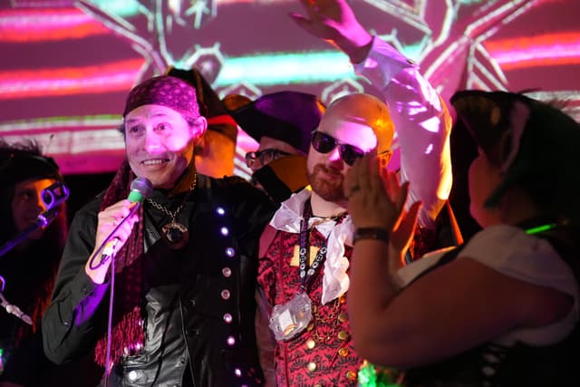 A group of people dressed in elaborate, colorful costumes, with one person holding a microphone and others engaging in lively interaction against a vibrant, illuminated backdrop