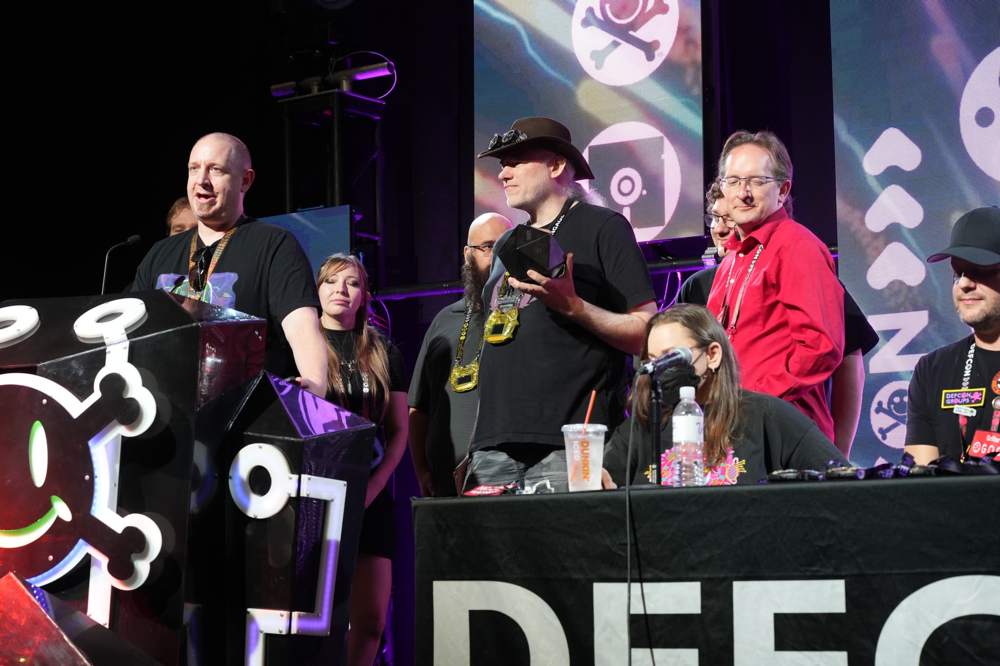 A group of people standing on a stage, some speaking into microphones, with a large banner displaying the word DEFCON in the foreground. The background features various symbols and designs