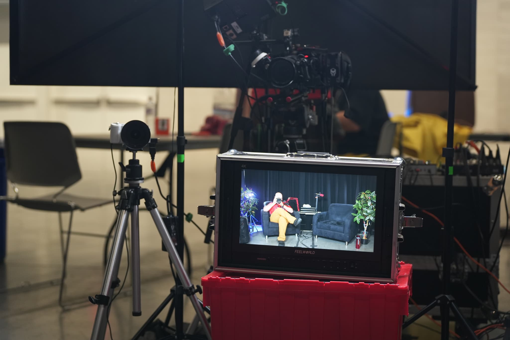 A video monitor displays a person sitting on a yellow chair in a studio setting, surrounded by various filming equipment including a camera on a tripod and lighting gear