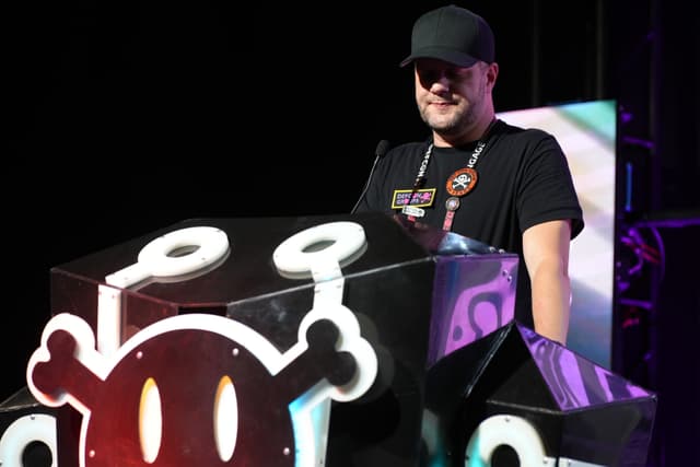 A person wearing a black cap and black t-shirt stands behind a DJ booth with a cartoonish skull and crossbones design