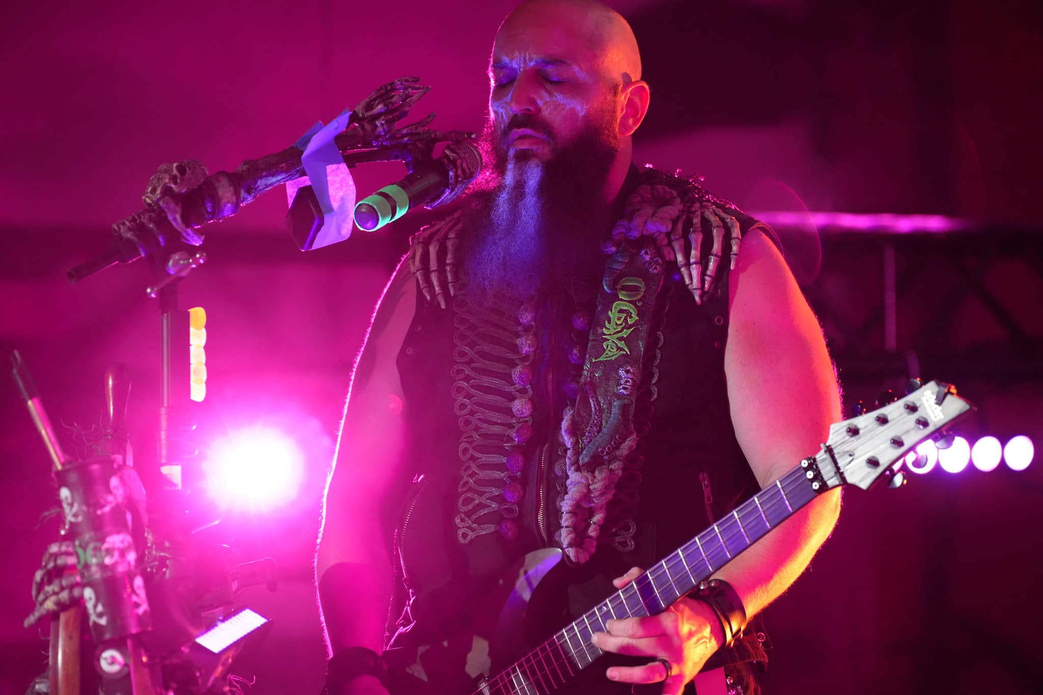 A musician with a long beard and a sleeveless outfit playing an electric guitar on stage, illuminated by vibrant pink and purple lights