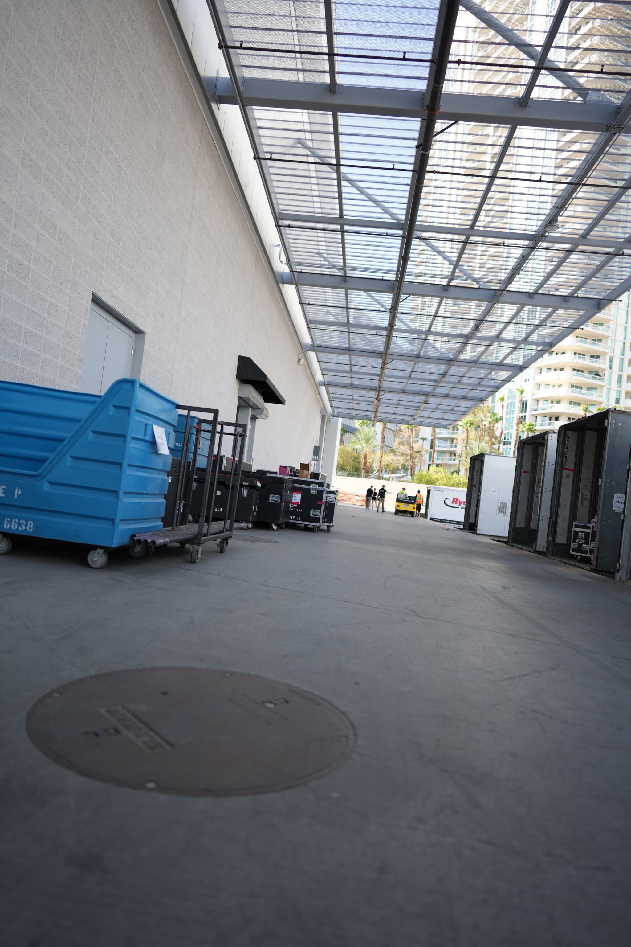 An industrial area with a covered walkway, blue dumpsters, and various equipment lined up against a white building