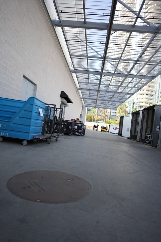 An industrial area with a covered walkway, blue dumpsters, and various equipment lined up against a white building