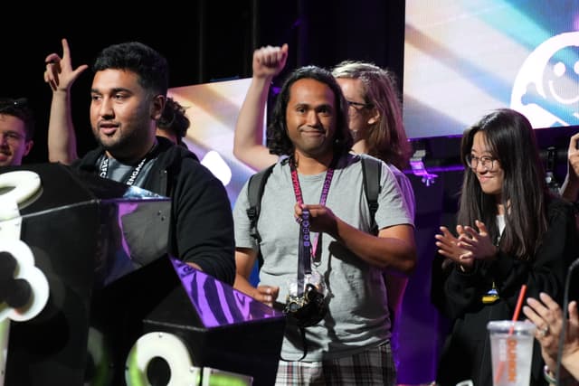 A group of people standing together, some clapping and others raising their fists in celebration, with a colorful background and stage lighting