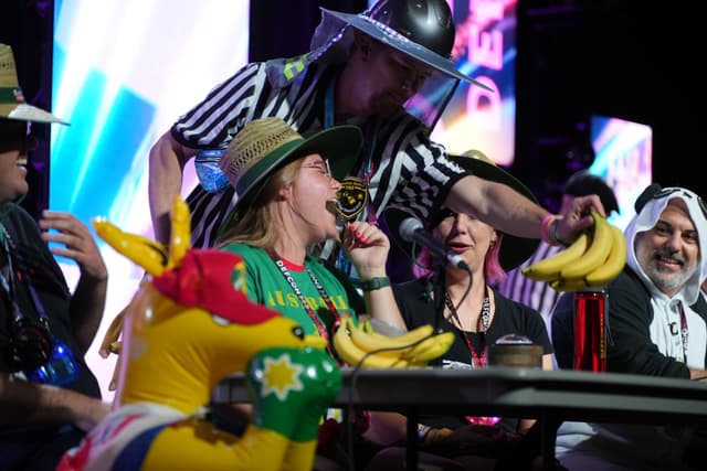 A group of people in colorful costumes and hats are gathered around a table, with one person enthusiastically eating a banana while another person, dressed as a referee, reaches over. Inflatable bananas and other props are also visible on the table