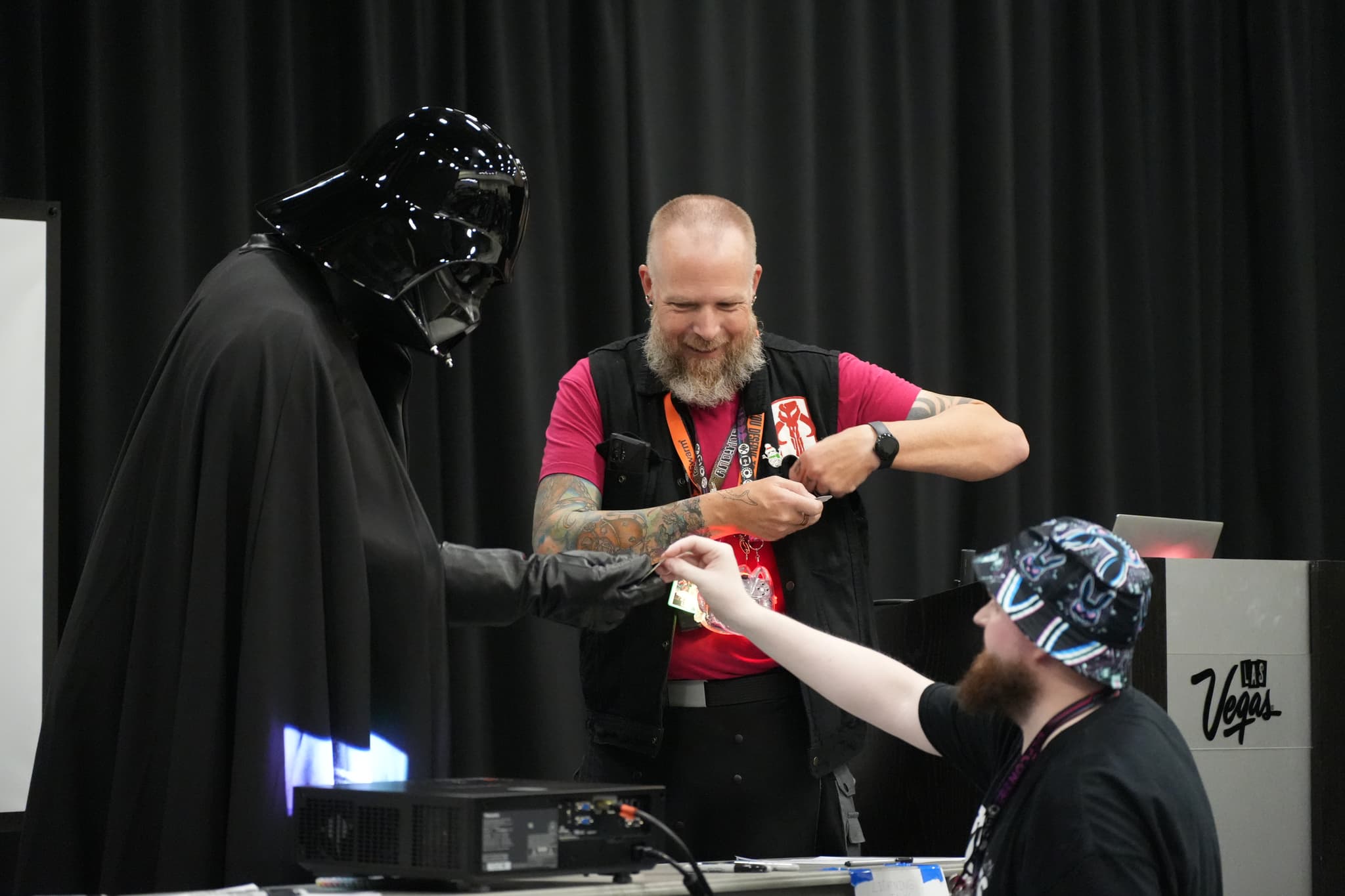 A person dressed as Darth Vader is interacting with two men, one of whom is handing something to the other