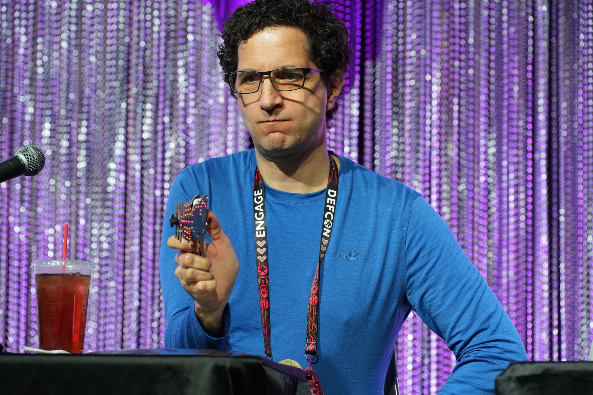 A man in a blue shirt and glasses is sitting at a table with a microphone and a drink, holding a small object in his hand, against a backdrop of shimmering purple curtains