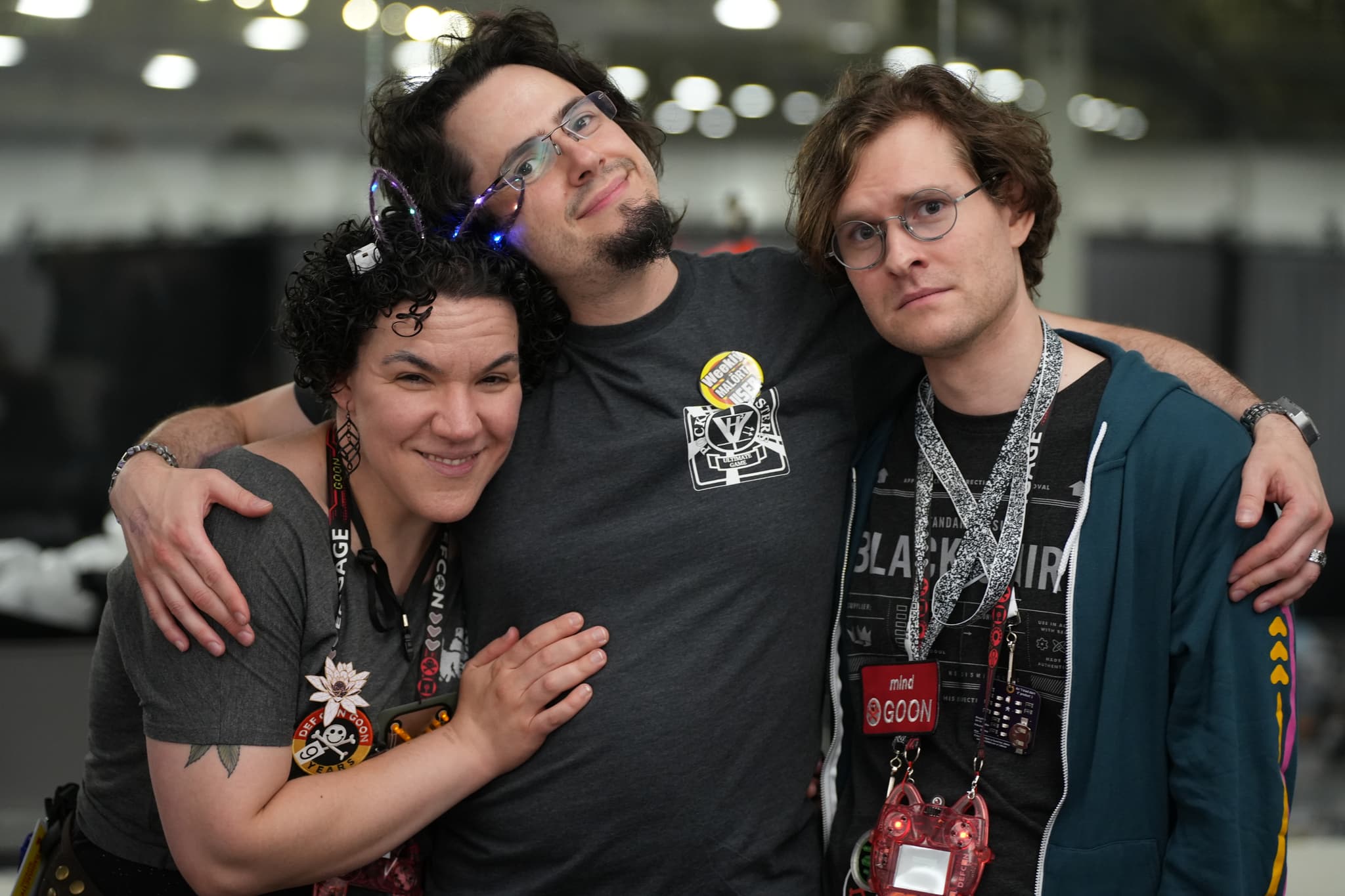 Three people standing close together, smiling, and wearing lanyards with badges. The person on the left has short curly hair and is wearing a headband with lights. The person in the middle has glasses and a beard, and the person on the right has glasses and wavy hair. The background is blurred, indicating an indoor setting with lights