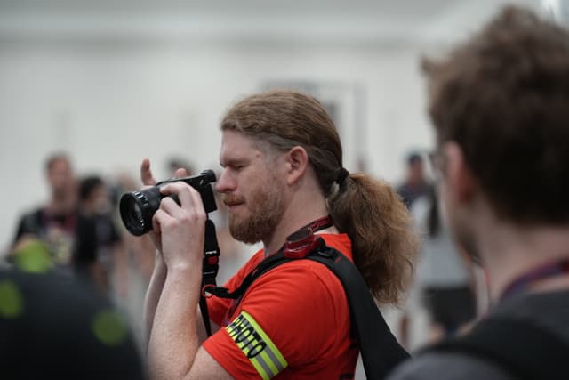 A person with long hair tied back, wearing a red shirt and a yellow armband, is taking a photo with a camera. Other people are blurred in the background