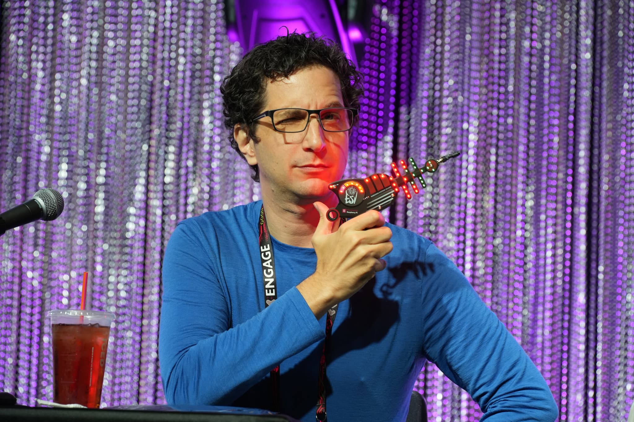 A person wearing glasses and a blue shirt holds a toy ray gun while sitting at a table with a microphone and a drink, against a backdrop of shimmering curtains