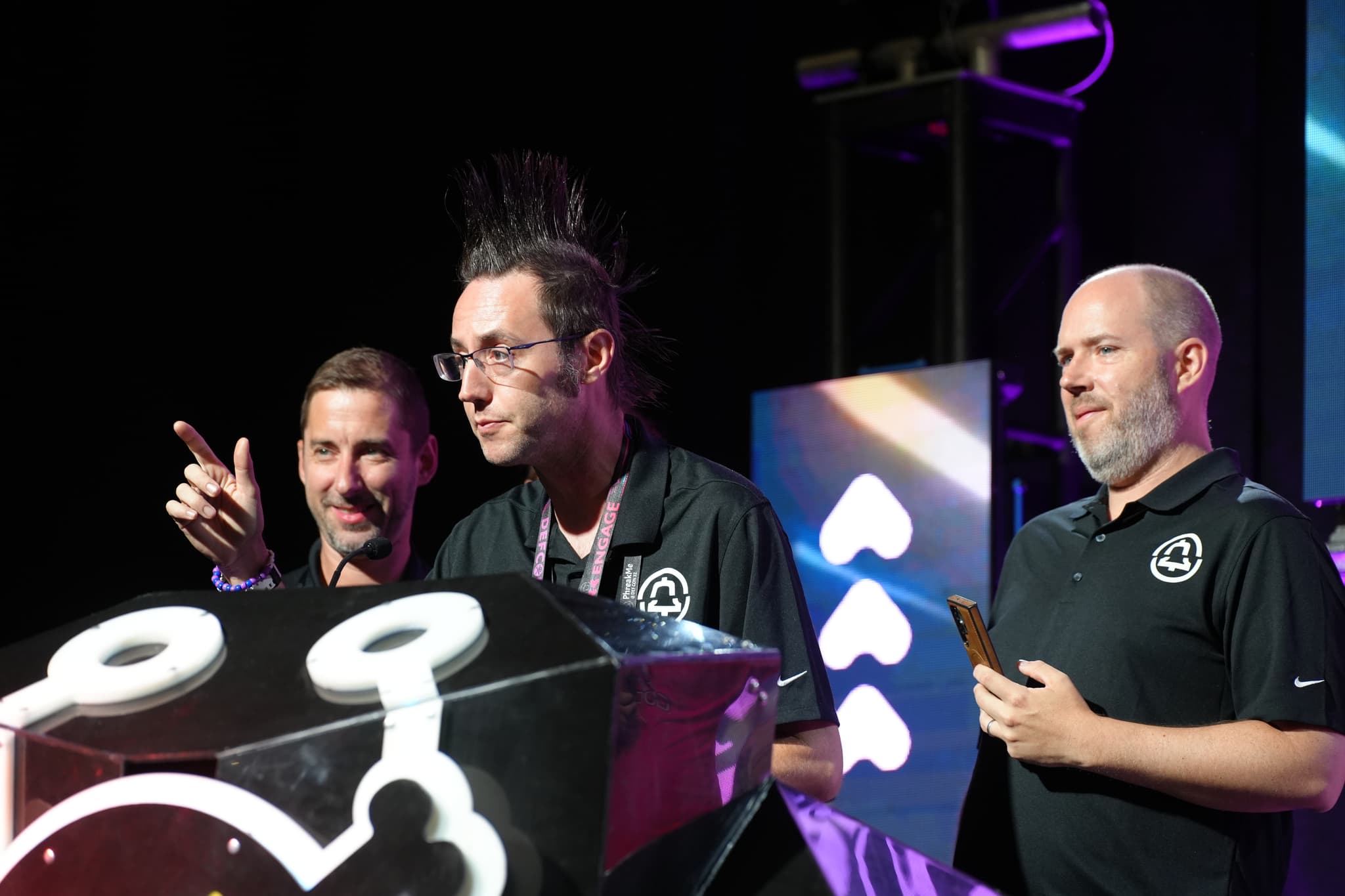 Three men standing on a stage, with one of them pointing and another holding a phone. They are behind a podium with a stylized design, and the background features illuminated symbols