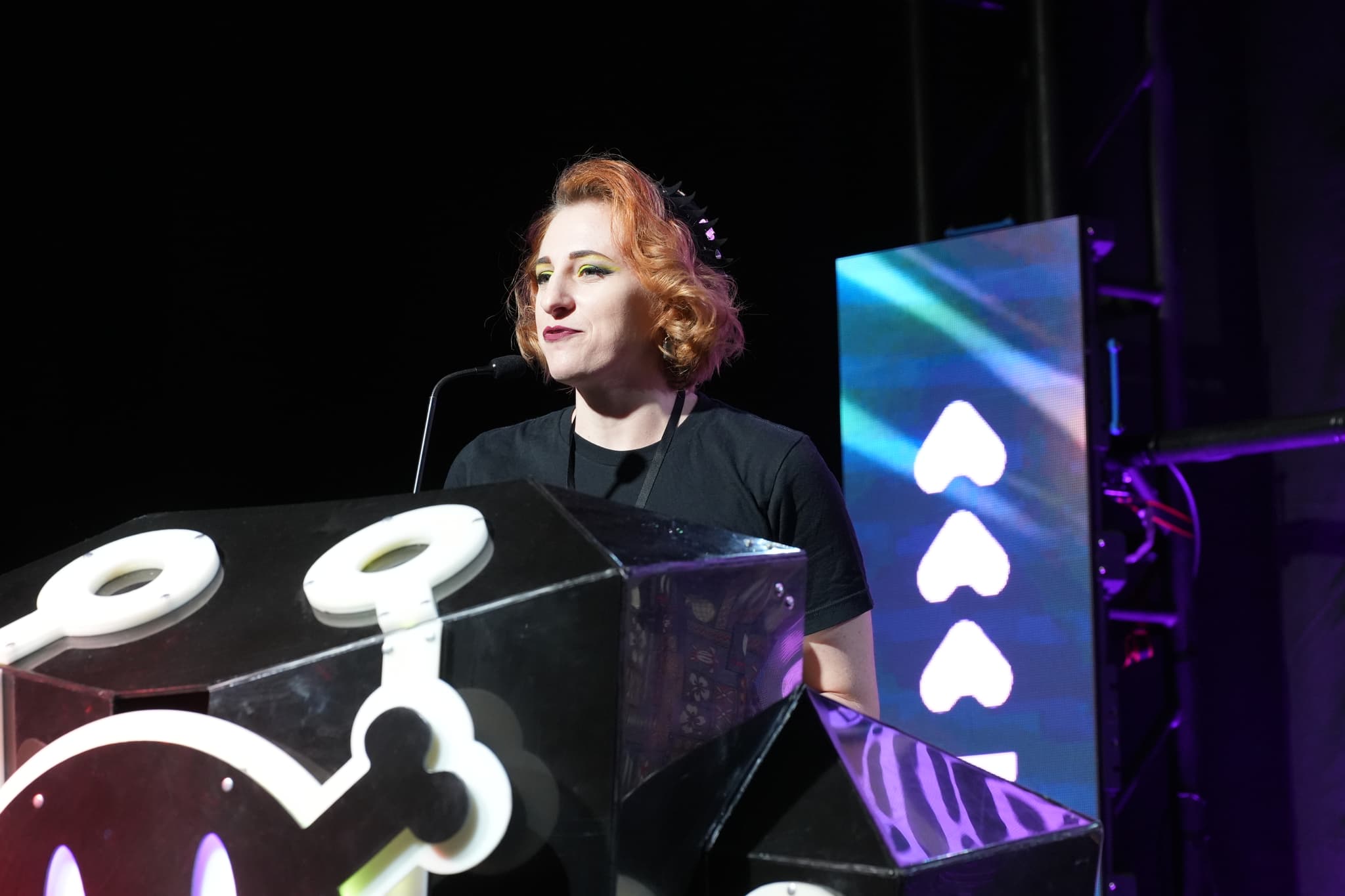 A person with short, reddish hair stands behind a podium with a microphone, illuminated by stage lights. The background features a screen with a pattern of white hearts on a blue and purple gradient