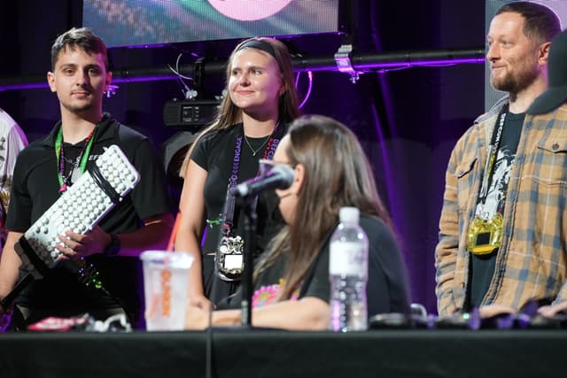 Four people on a stage, with one person holding a keyboard and another speaking into a microphone