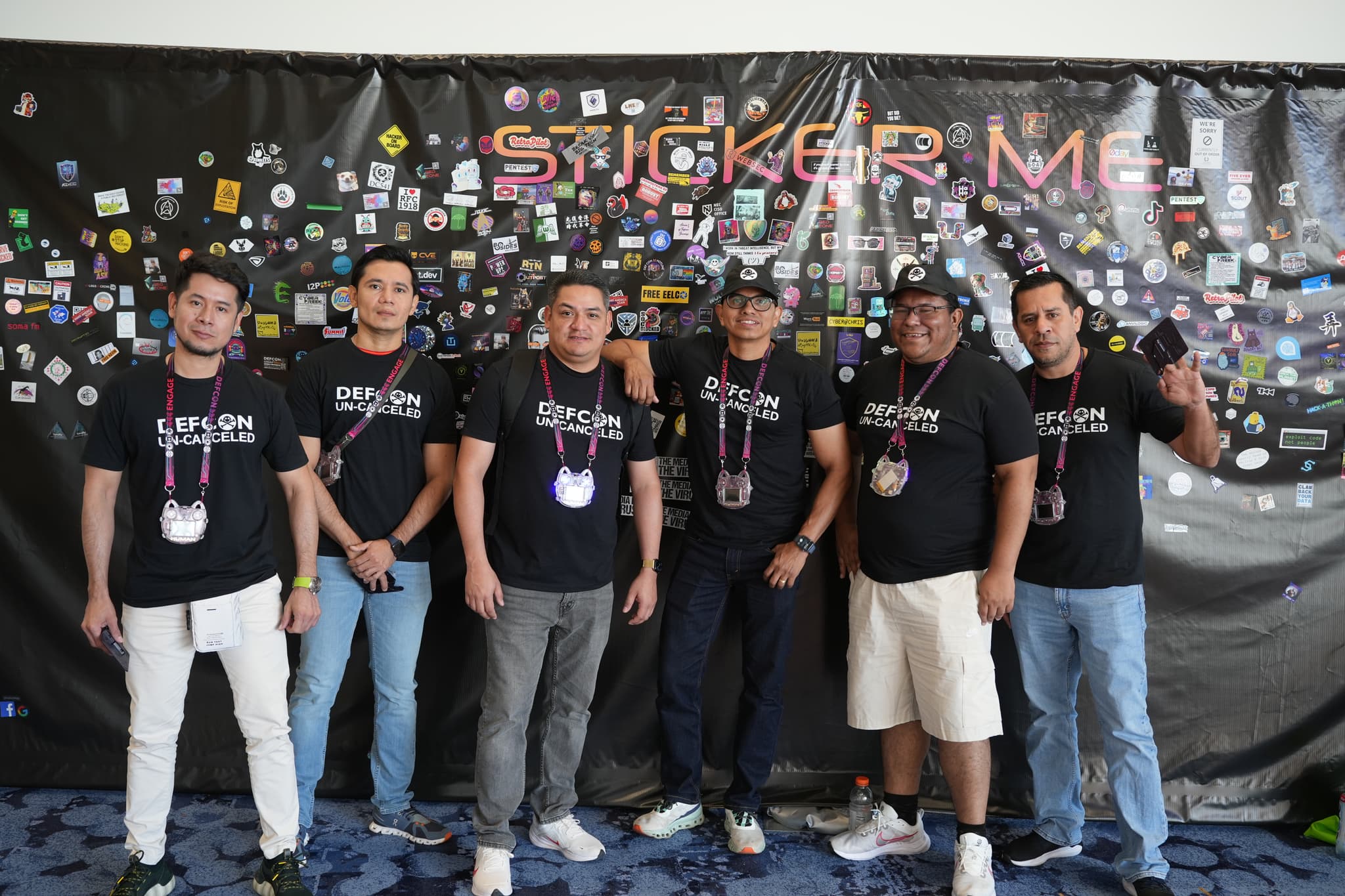 Six men standing in front of a black backdrop covered with various stickers, all wearing matching black t-shirts and lanyards