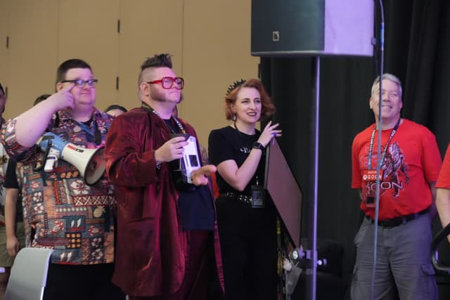 A group of four people standing together, with one person holding a bottle and another person holding a microphone. They appear to be at an indoor event or gathering