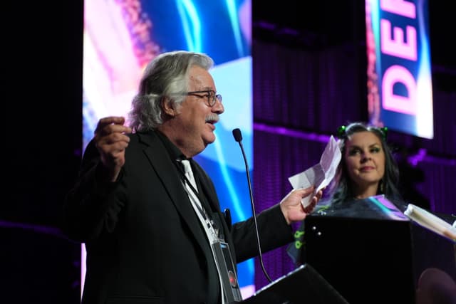 An older man in a suit speaks at a podium, holding papers, while a woman stands nearby, looking on