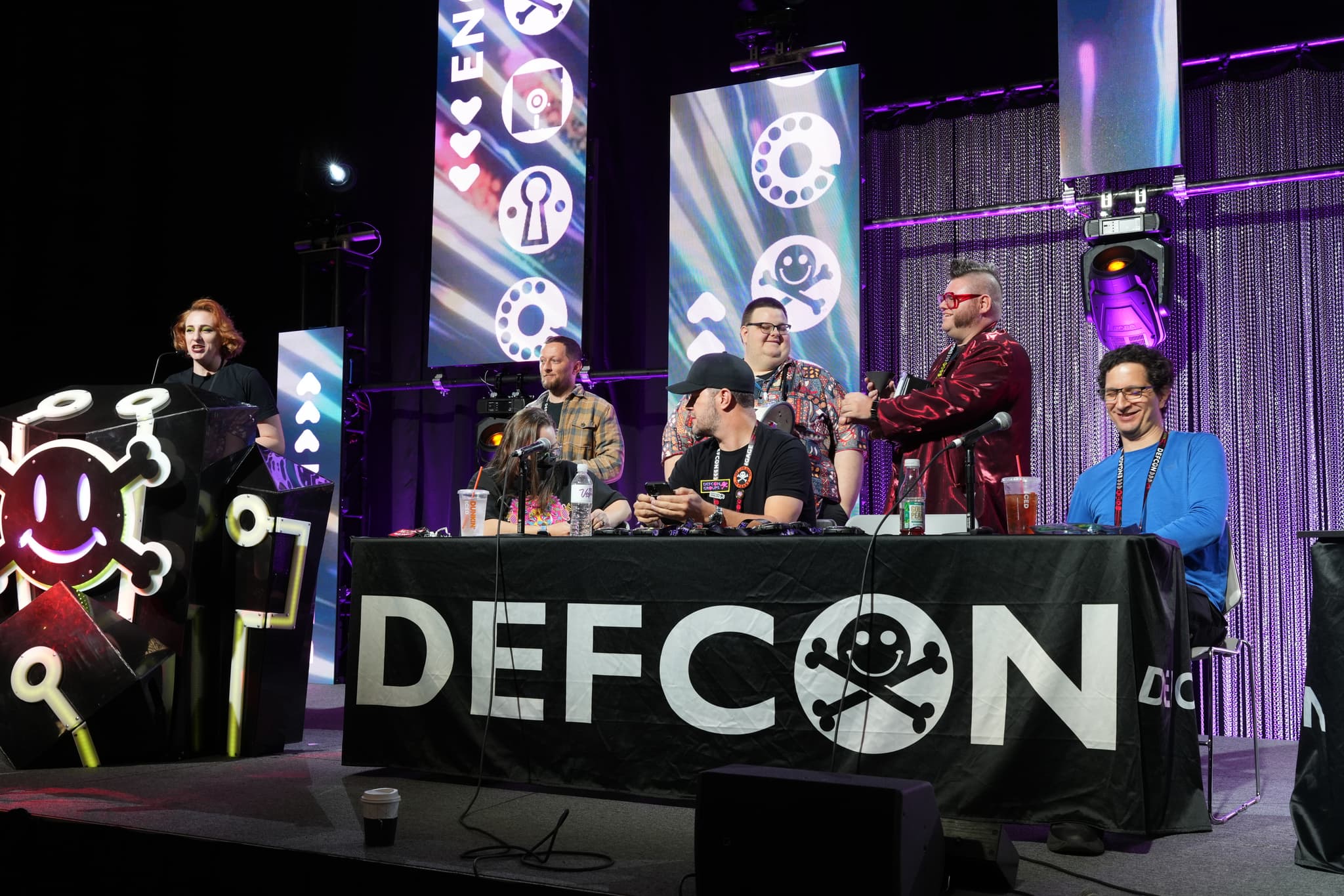 A panel of six individuals seated at a table with a DEFCON banner, featuring a skull and crossbones logo. The background includes screens displaying various symbols and a purple curtain. One person is standing at a podium on the left side