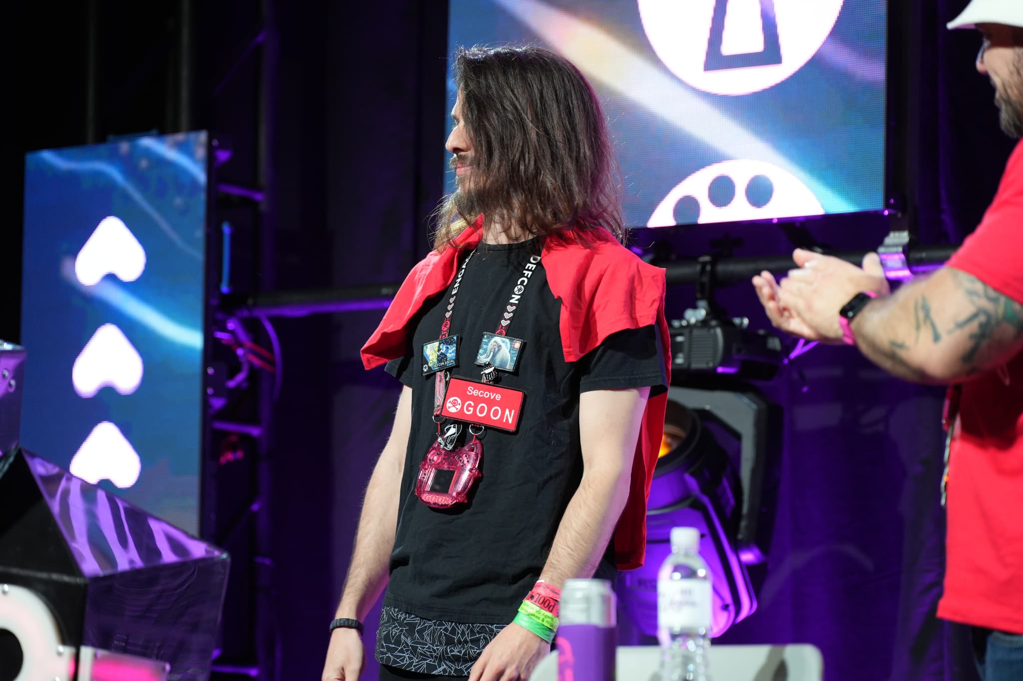 Two individuals are on a stage with colorful lighting and screens in the background. One person, with long hair, is wearing a black t-shirt with a red garment draped over their shoulders and a lanyard around their neck. The other person, wearing a red shirt and cap, is clapping. Various objects, including water bottles, are on a table in front of them