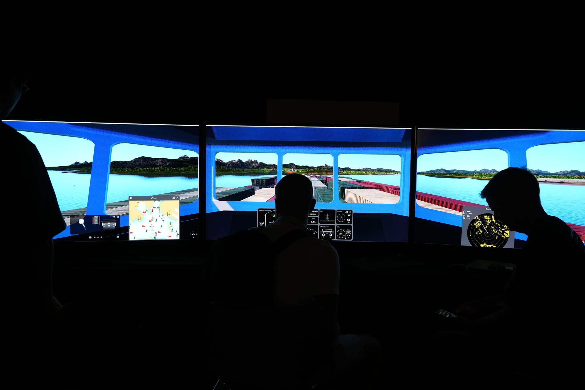 Three people in a dark room operating a boat simulator with multiple screens displaying a waterway