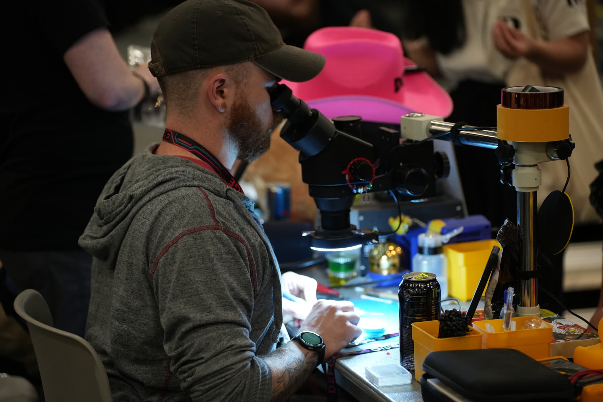 A person wearing a cap and hoodie is working at a cluttered desk with a microscope and various tools, with a bright pink hat in the background