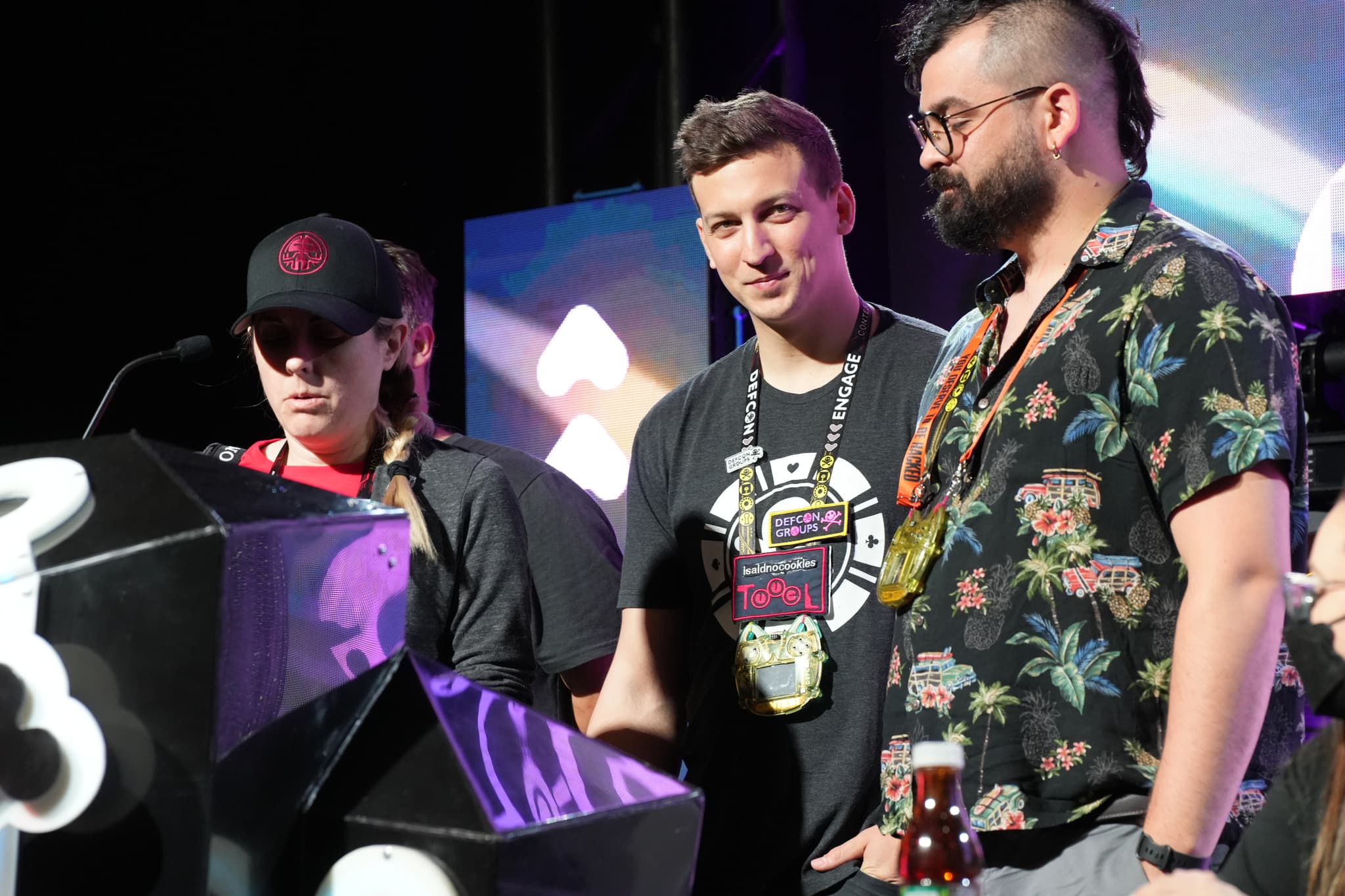 Three individuals standing on a stage, with one speaking into a microphone. They are dressed casually, and the background features colorful lighting and geometric shapes