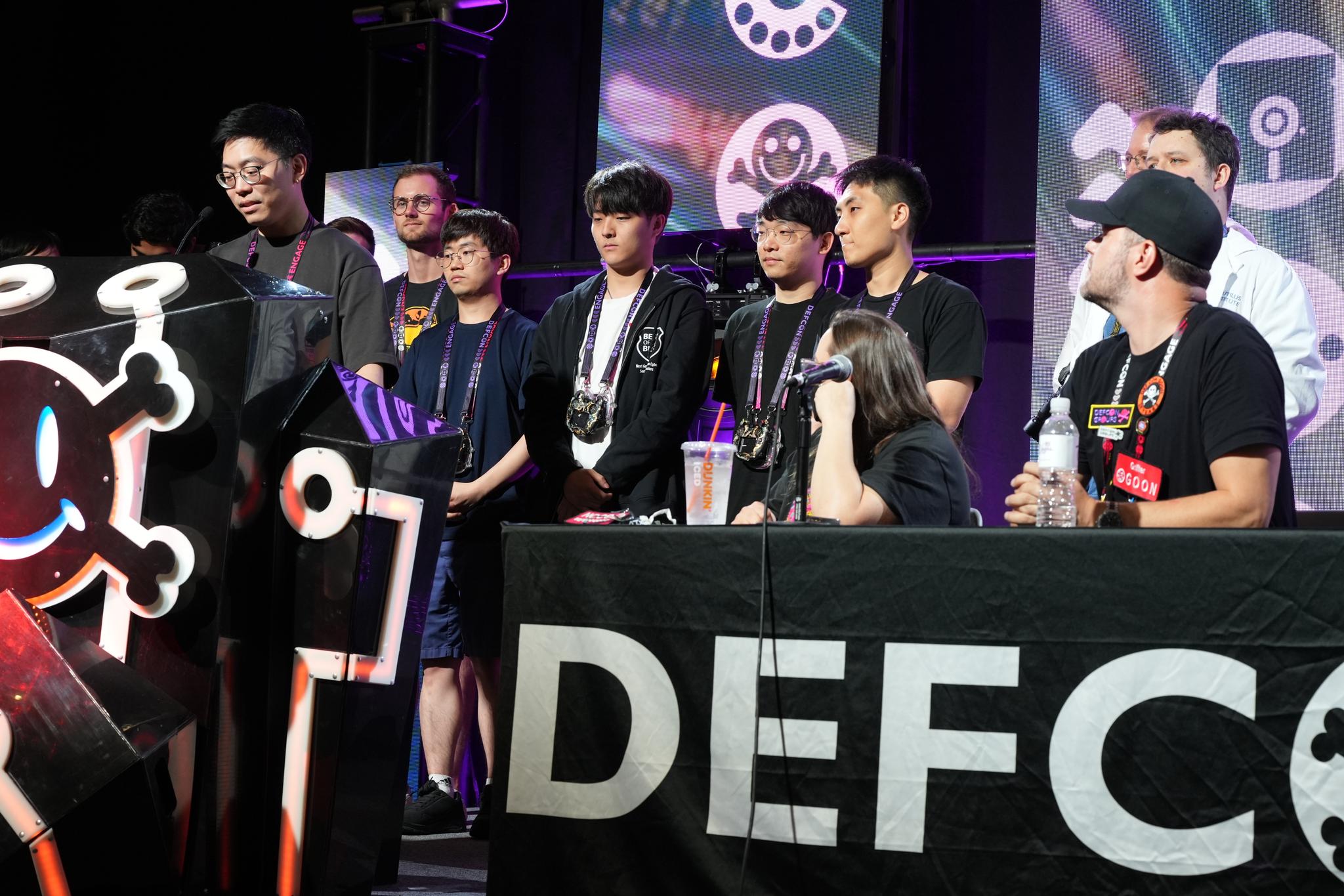 A group of people on stage at DEFCON, with some standing and others sitting behind a table. The stage features a large DEFCON banner and various electronic equipment