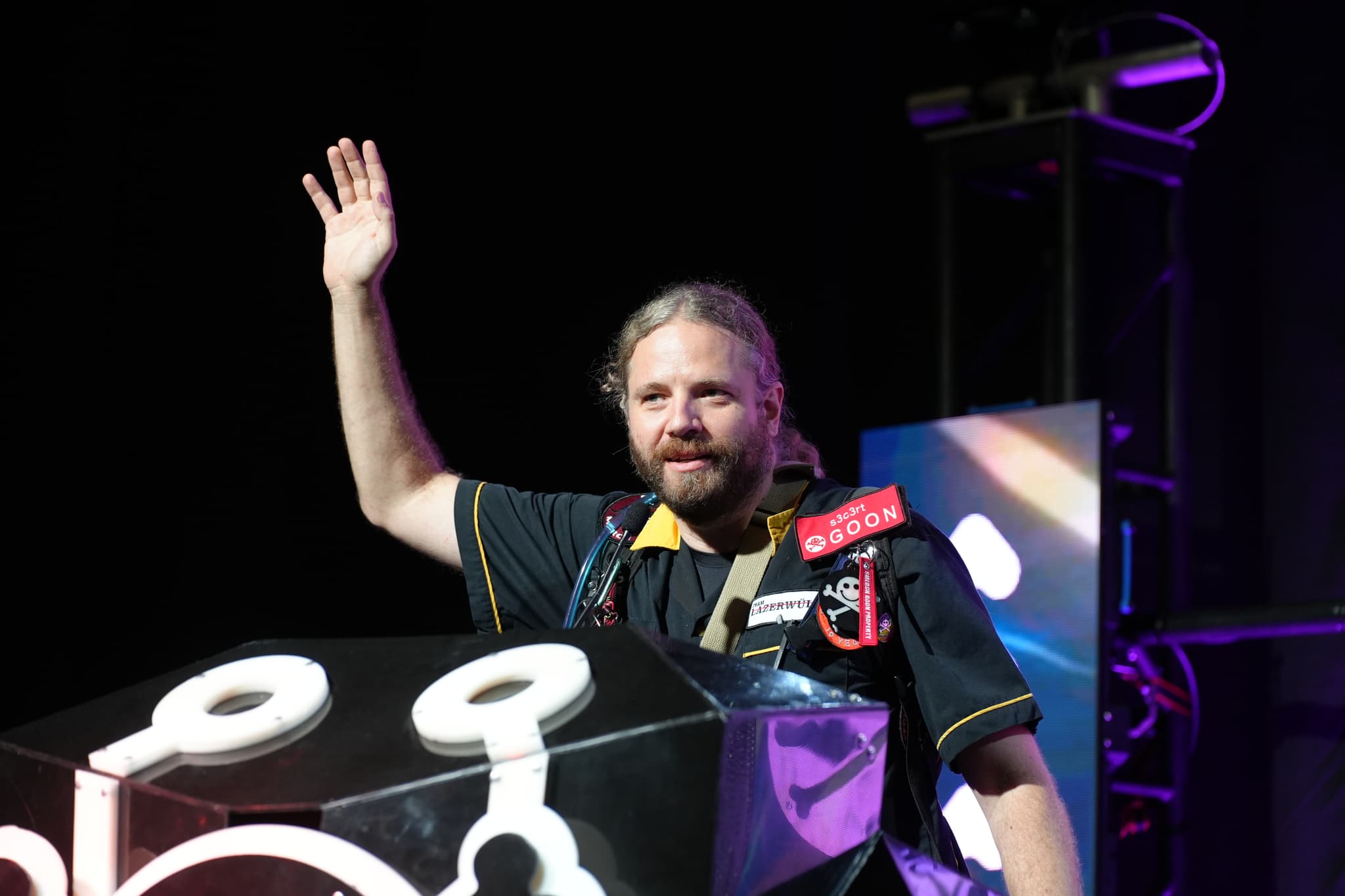 A person with long hair and a beard, wearing a dark shirt, stands behind a podium with a raised hand. The background is dark with some illuminated elements