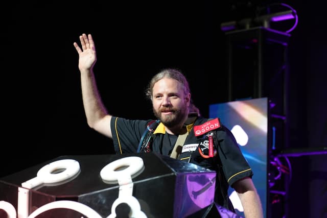 A person with long hair and a beard, wearing a dark shirt, stands behind a podium with a raised hand. The background is dark with some illuminated elements