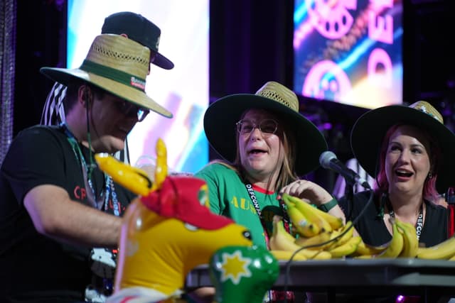 Three people wearing hats are gathered around a table with inflatable kangaroos and bananas, appearing to be engaged in a lively and festive activity. The background features colorful lights and decorations