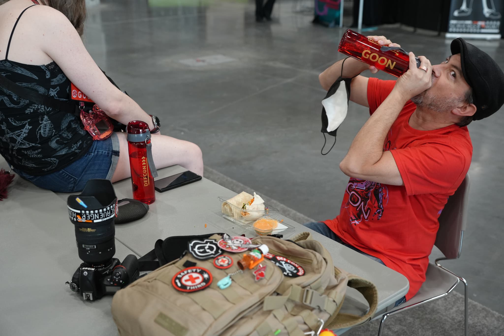 Two people sitting at a table, one drinking from a large red bottle and the other holding a similar bottle. The table has a camera, a bag with various pins and patches, and some food items