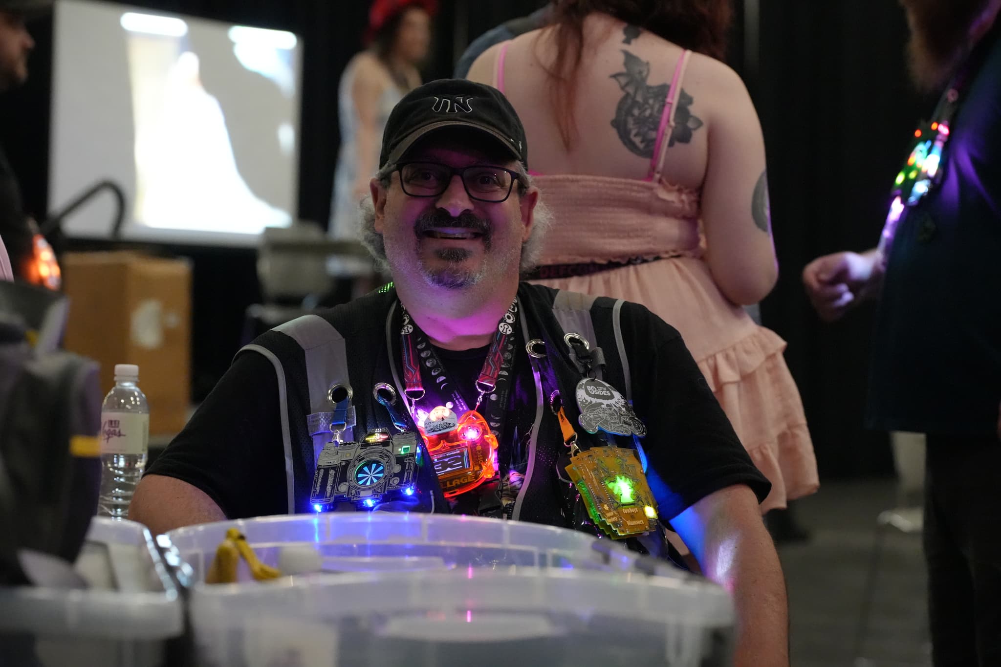 A person wearing a cap and a vest adorned with colorful, glowing lights is smiling while sitting at a table. Other people and a screen are visible in the background