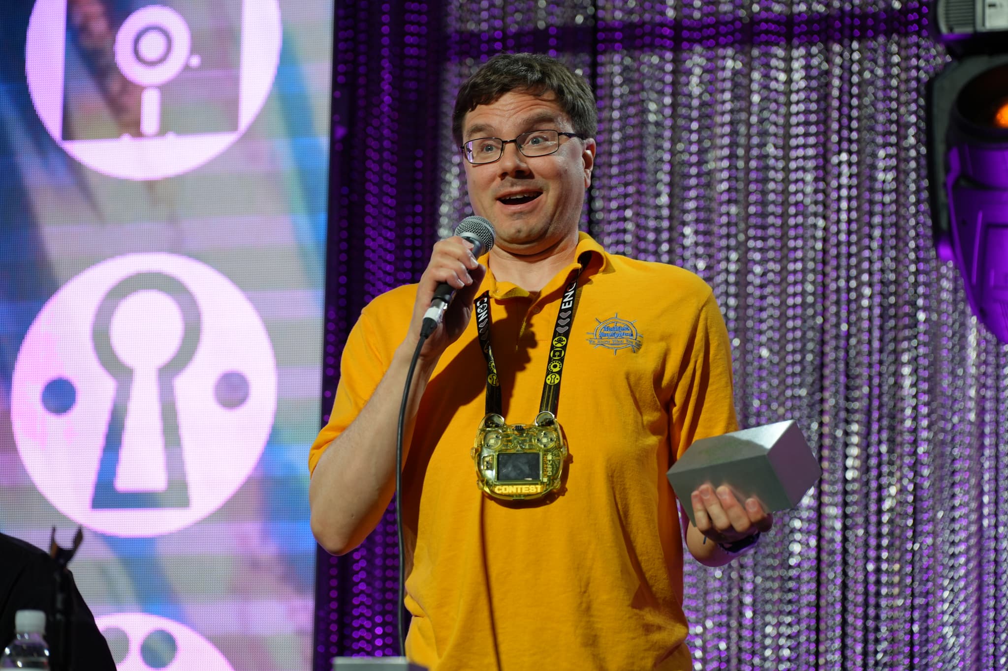 A man wearing a yellow shirt and a large lanyard is speaking into a microphone while holding a piece of paper. The background features a curtain with a shimmering texture and circular designs with keyhole symbols