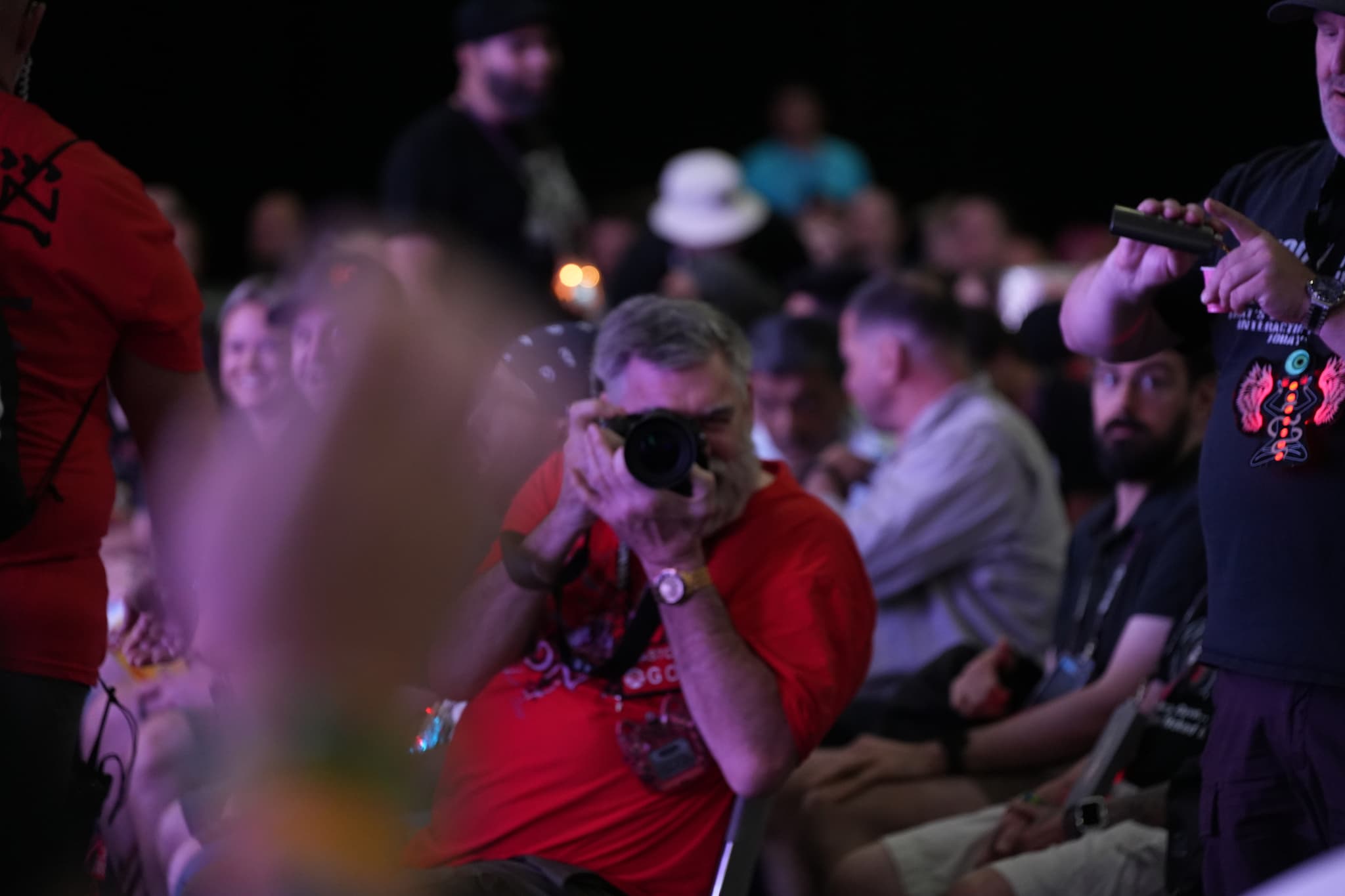 A person in a red shirt is taking a photo with a camera in a crowded indoor setting, surrounded by people who appear to be engaged in an event or gathering