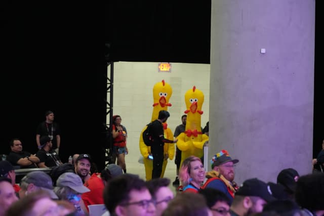 A crowd of people, with two individuals in large yellow chicken costumes entering through a doorway in the background