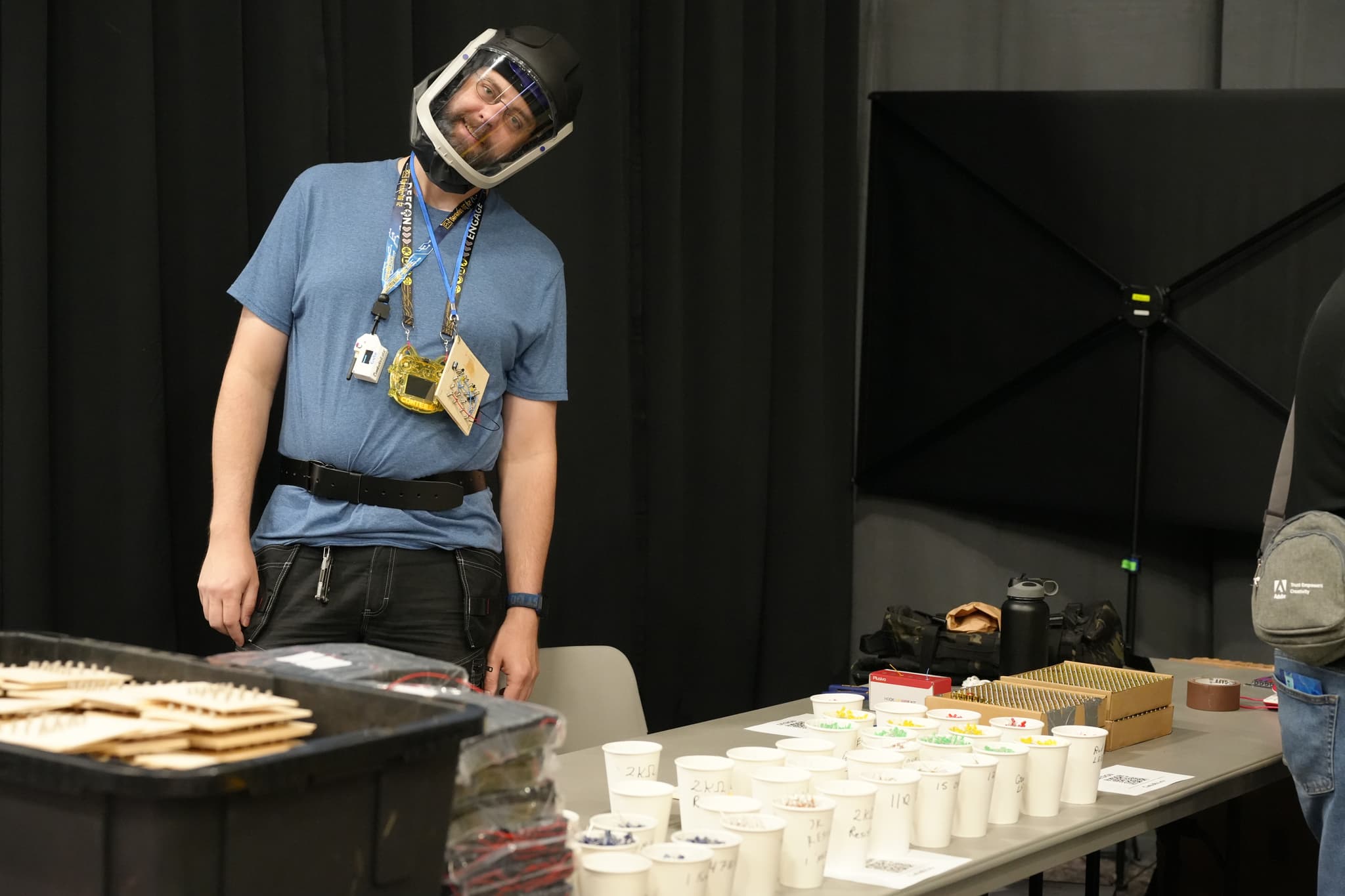 A person wearing a helmet and a blue shirt stands next to a table with various items, including cups and containers, in a room with black curtains