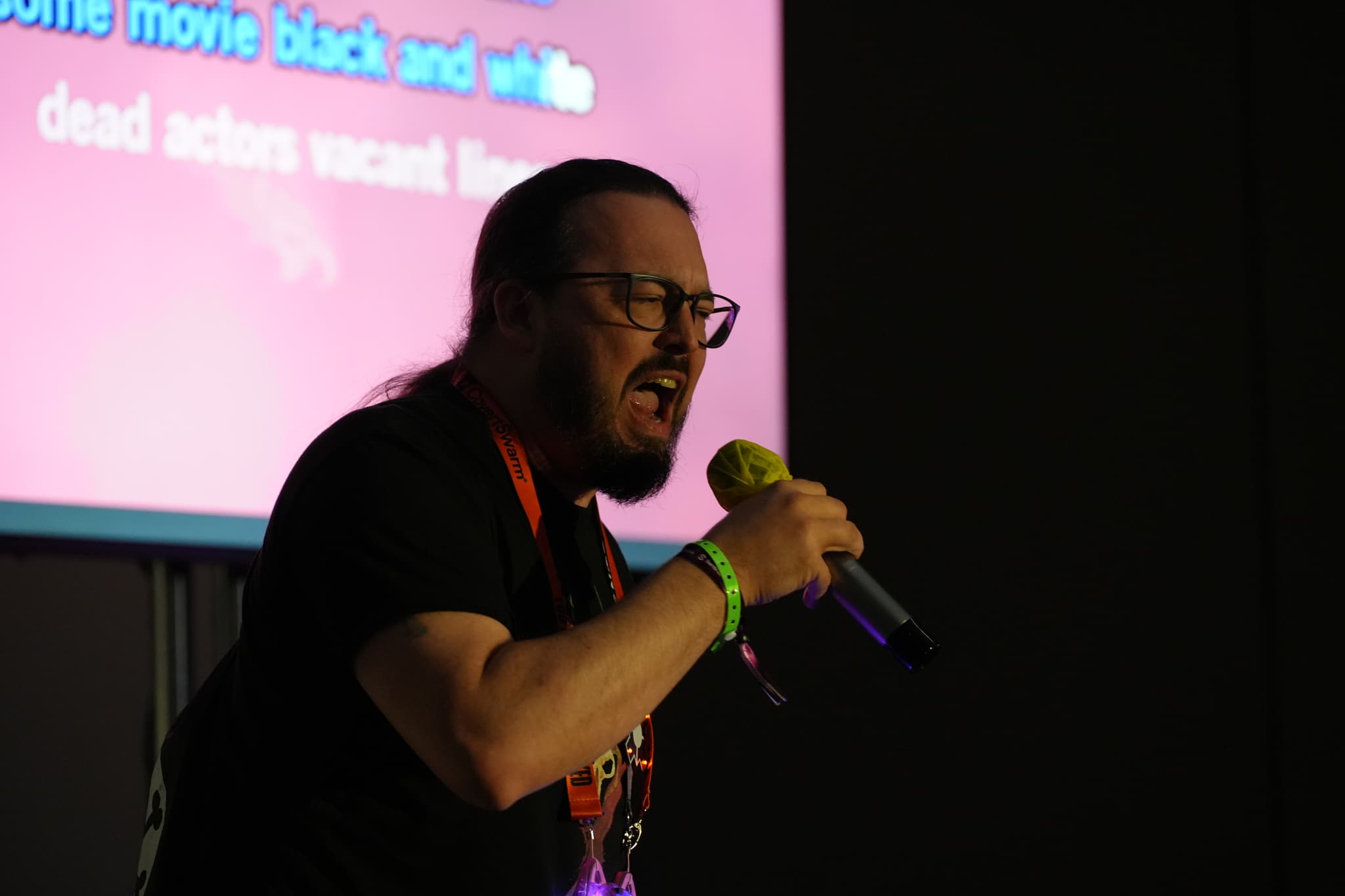 A person with long hair and glasses is passionately singing into a microphone, with a colorful screen displaying text in the background