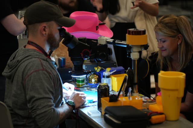 Two people working at a cluttered table with various tools and equipment, including a microscope and yellow containers, while others are visible in the background